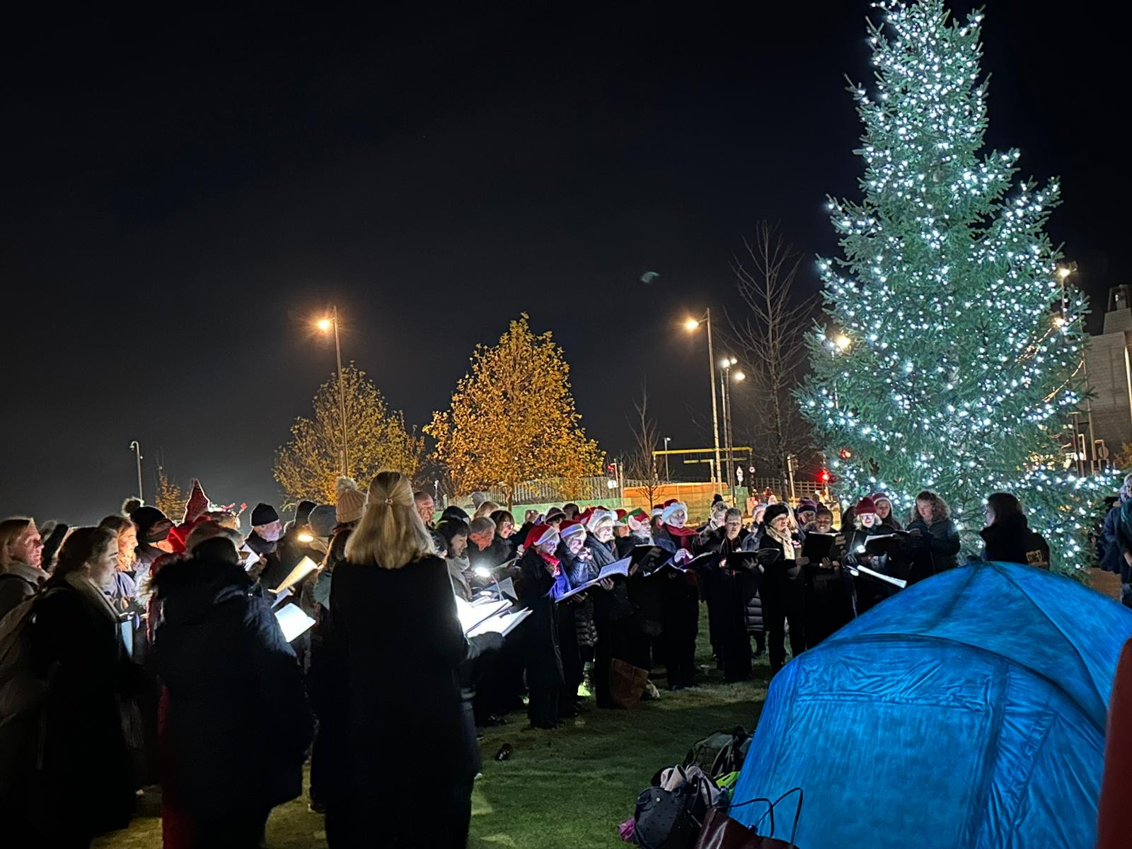 Carollers singing on The Green, the sky is lit up by the large sparkling Campus Christmas tree.